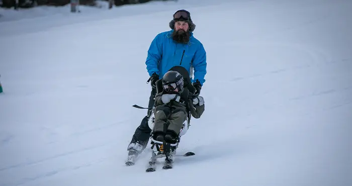 Man åker skidor och ett barn sitter på en anpassad skida.