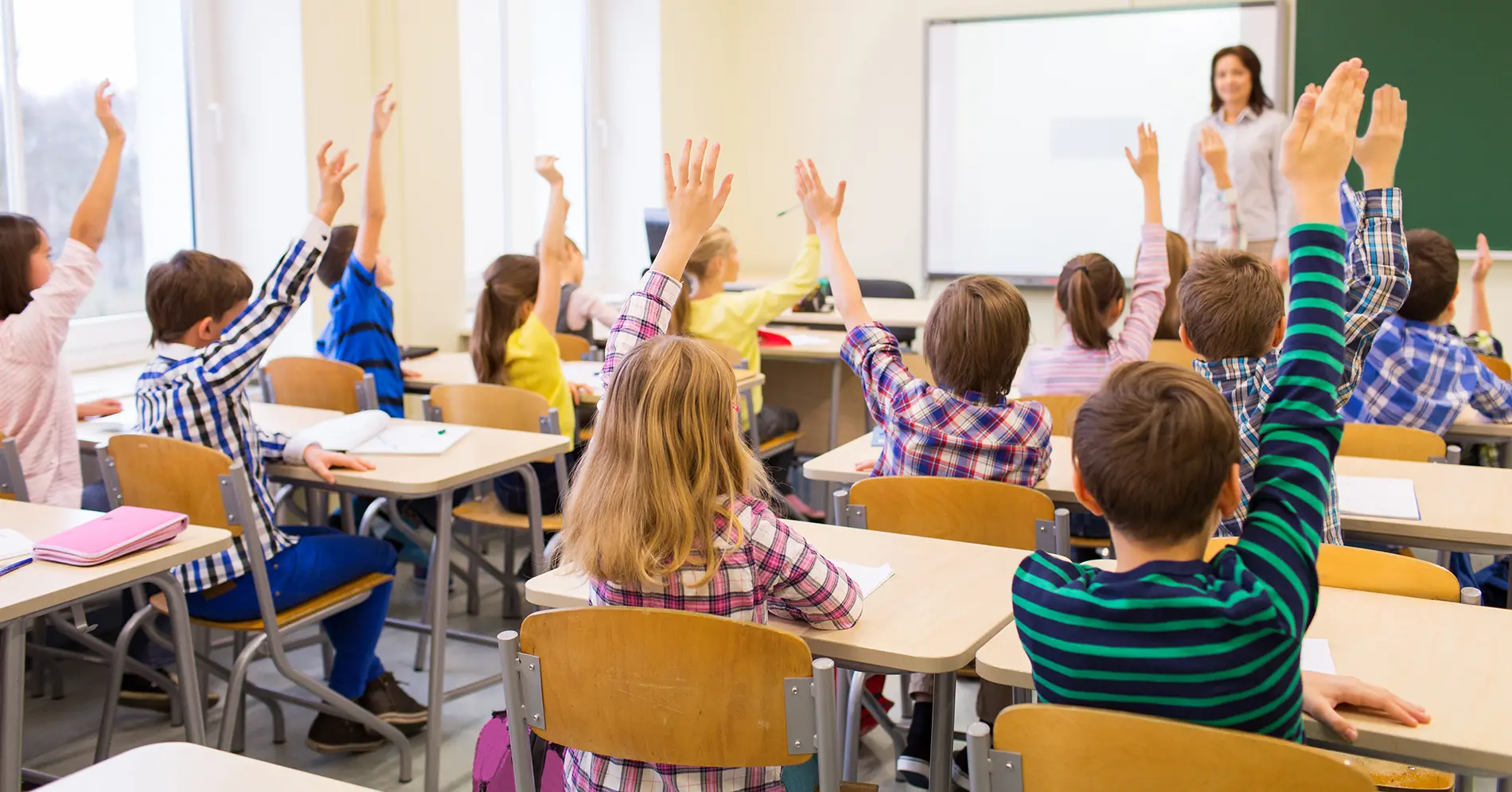 Ett klassrum med barn som sitter i bänkar och räcker upp handen.